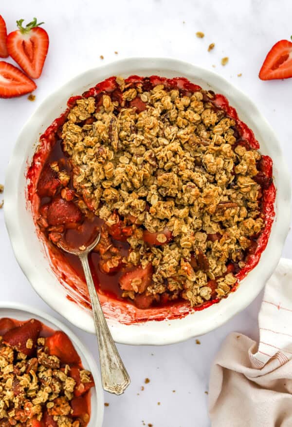 Pie dish filled with a healthy strawberry crumble with more sliced strawberries behind it and some crumble in a bowl in front of it.