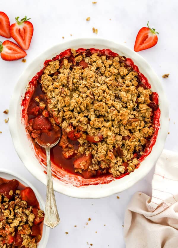 White pie dish filled with strawberry crumble with more in a bowl in front of it and sliced strawberries behind it.