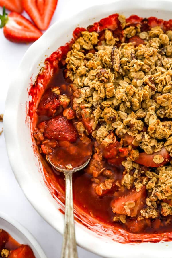 Baked strawberry crumble in a white pie dish with a spoon in the dish and sliced strawberries behind it.