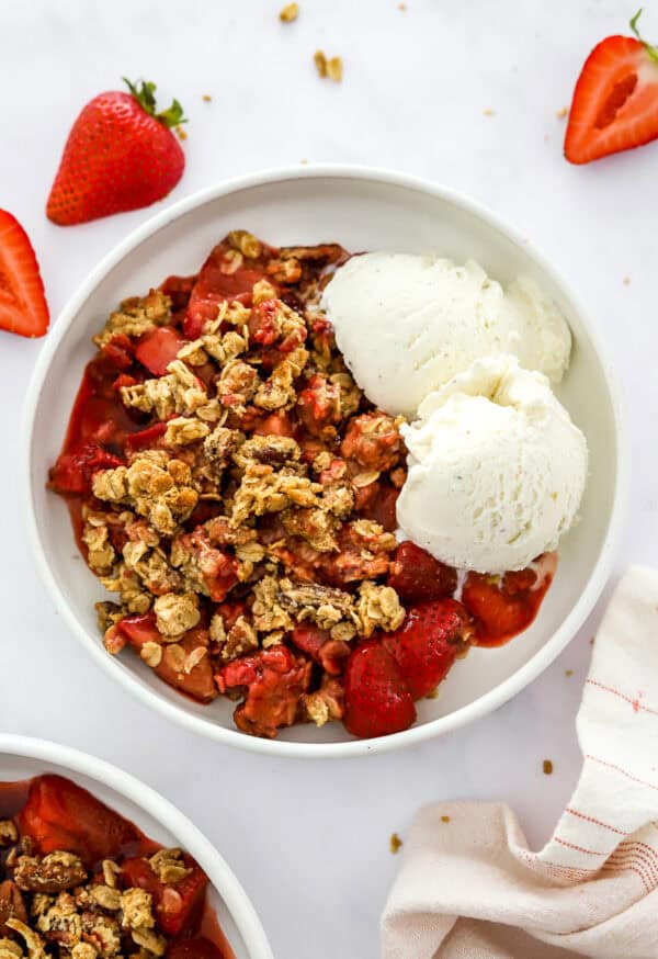 Bowl of strawberry crisp with vanilla ice cream with another bowl of it in front of it.
