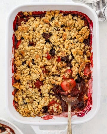 Baking dish filled with baked berry crisp with a spoon in the dish and more crisp in a bowl in front of it.