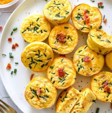 Plate of egg bites topped with chives and bacon with more cheese and chives behind it and a spoon and linen in front of it.
