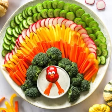 Round platter with a turkey veggie tray on it with a bowl of dip with a turkey face in it and some pumpkins and veggies around it.