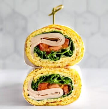 Two halves of an egg wrap turkey sandwich wrapped in parchment paper stacked on top of one another with a toothpick in the top.