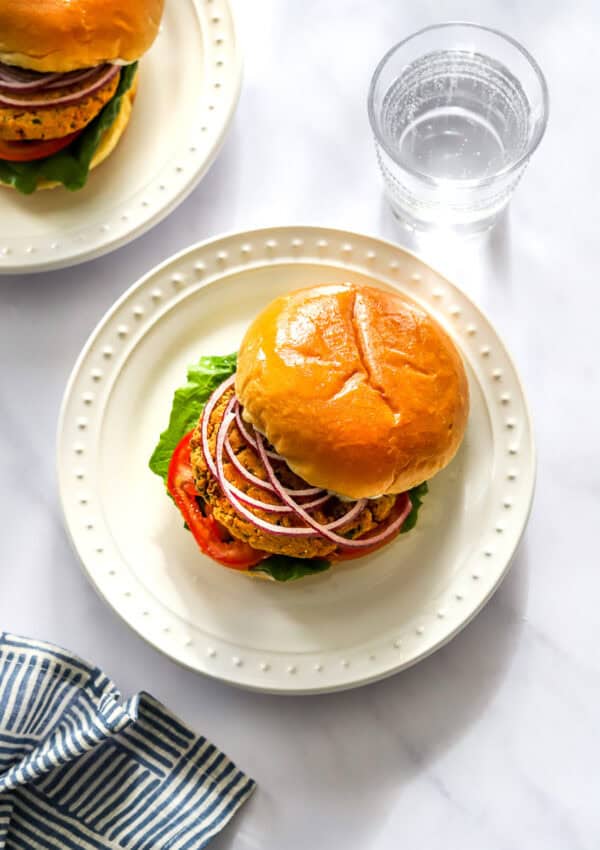 White plate with a chickpea cauliflower burger in a bun with sliced onion, lettuce and tomato on it with a glass of water and another burger on a plate behind it.