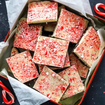 Red square tin filled with pieces of chocolate peppermint bark with candy canes and crushed peppermint around it.