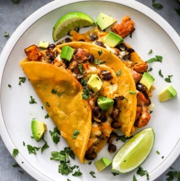 Round plate with two crispy black bean sweet potato tacos with cheese on it topped with diced avocado and cilantro with a lime wedge on the plate with it.