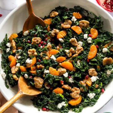 Kale pomegranate salad in a white salad bowl with wood serving spoon in the bowl and a cut pomegranate behind it.