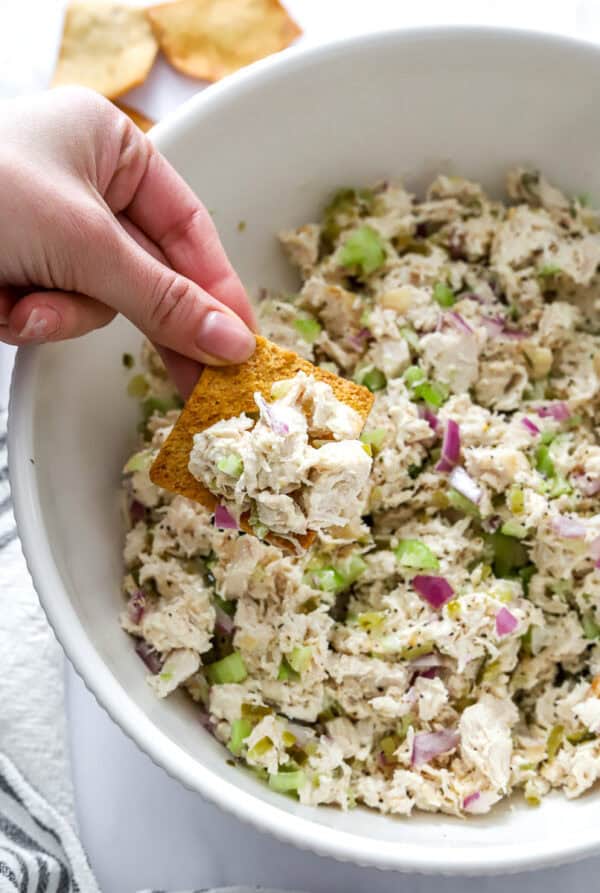 Hand lifting a chip from a bowl of blended chicken salad with a striped towel in front of it.