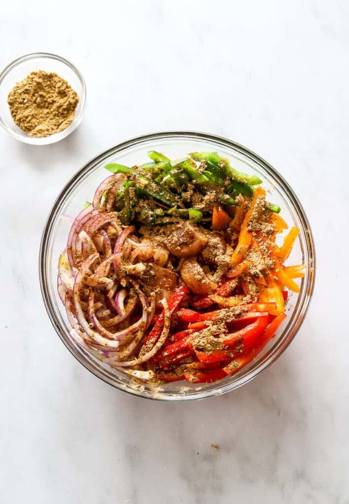 Sliced bell peppers, onion and chicken breast covered in oil and fajita seasoning in a glass bowl.