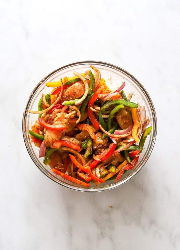 Seasoned, sliced uncooked peppers and chicken breast in a glass bowl.