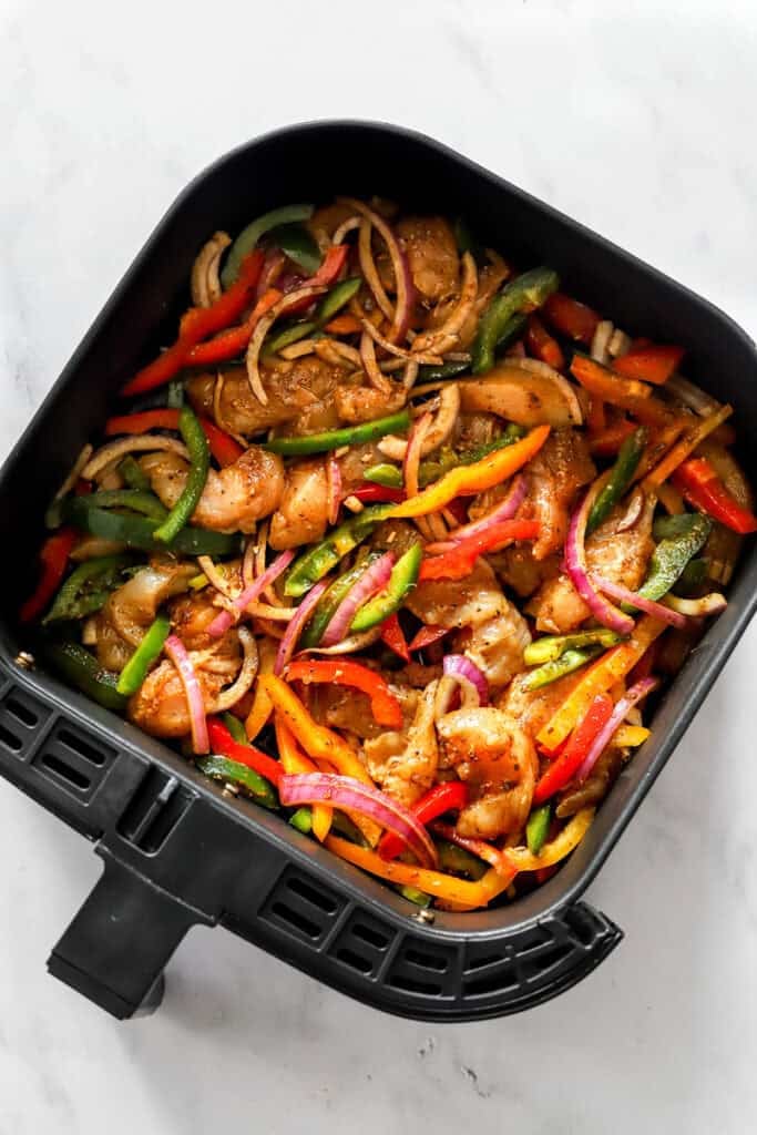 Seasoned, uncooked sliced peppers and chicken in a black air fryer basket.
