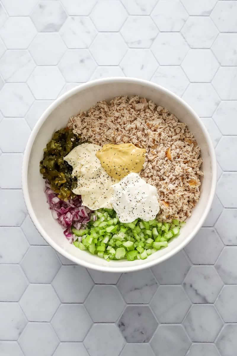 Shredded cooked chicken, chopped purple onion, diced celery, mayo, mustard, and Greek yogurt in a white mixing bowl before being mixed.