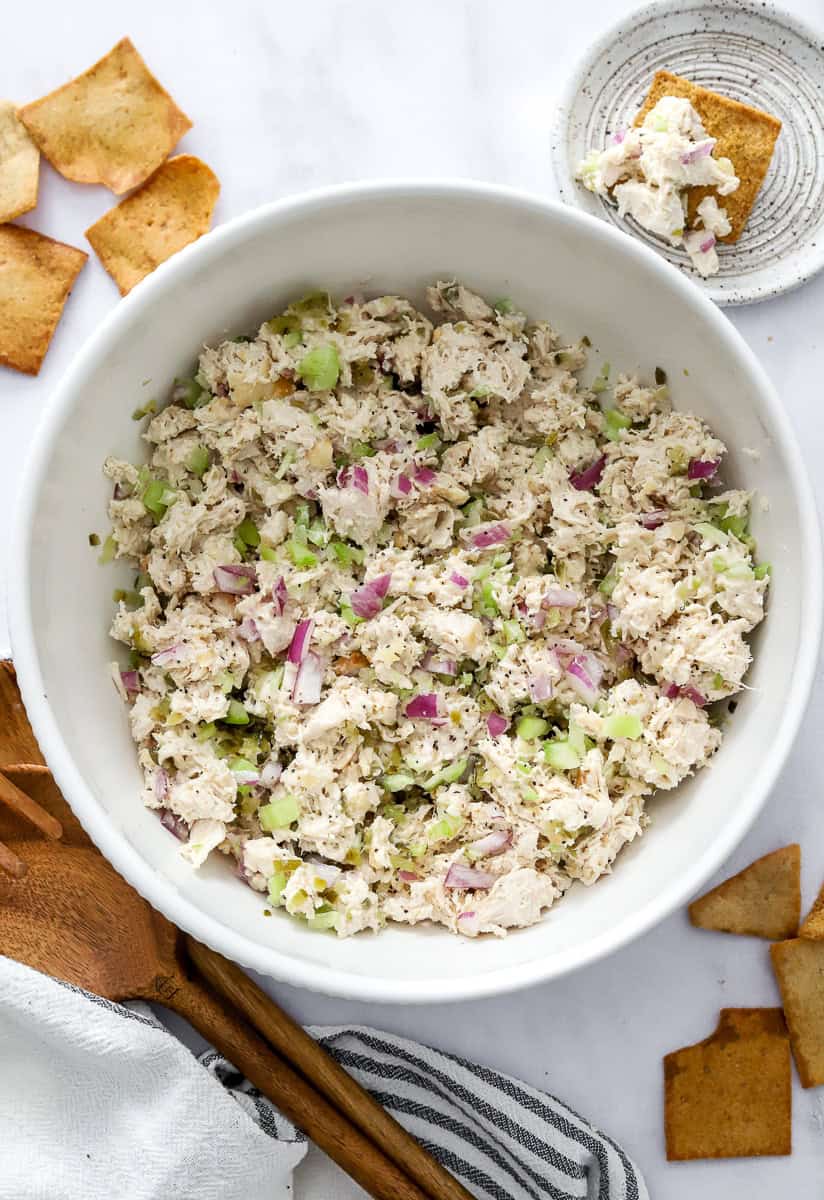 White serving bowl filled with mixed chicken salad with chopped onion and celery in it with wooden serving spoons and pita chips in front of it.
