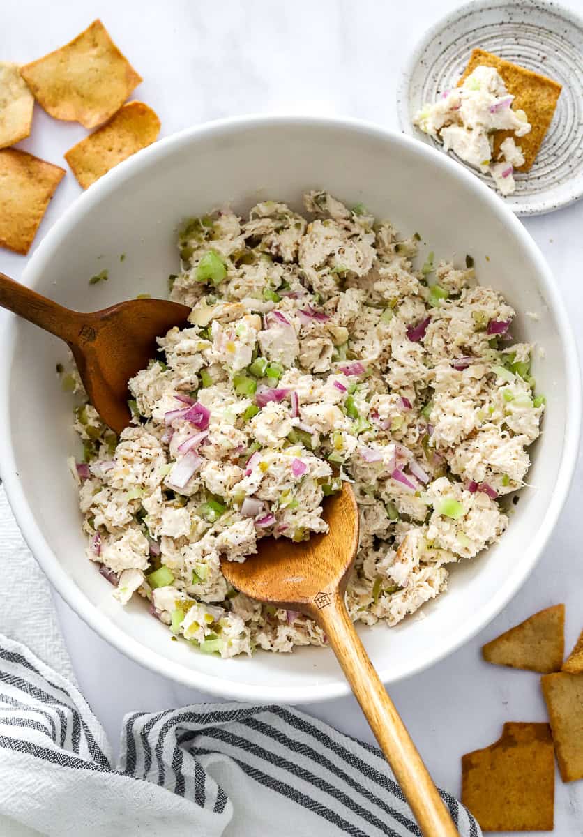 Serving spoons in a bowl of rotisserie chicken salad with pita chips around the bowl and a striped towel in front of it.
