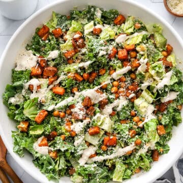 kale caesar salad in a white salad bowl with wood serving spoons next to it