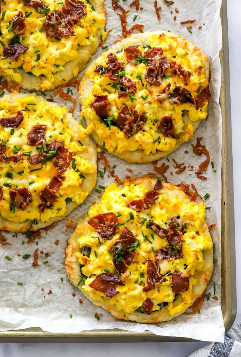 4 baked breakfast style pizzas on a lined pan with scrambled eggs, bacon and chives on them.