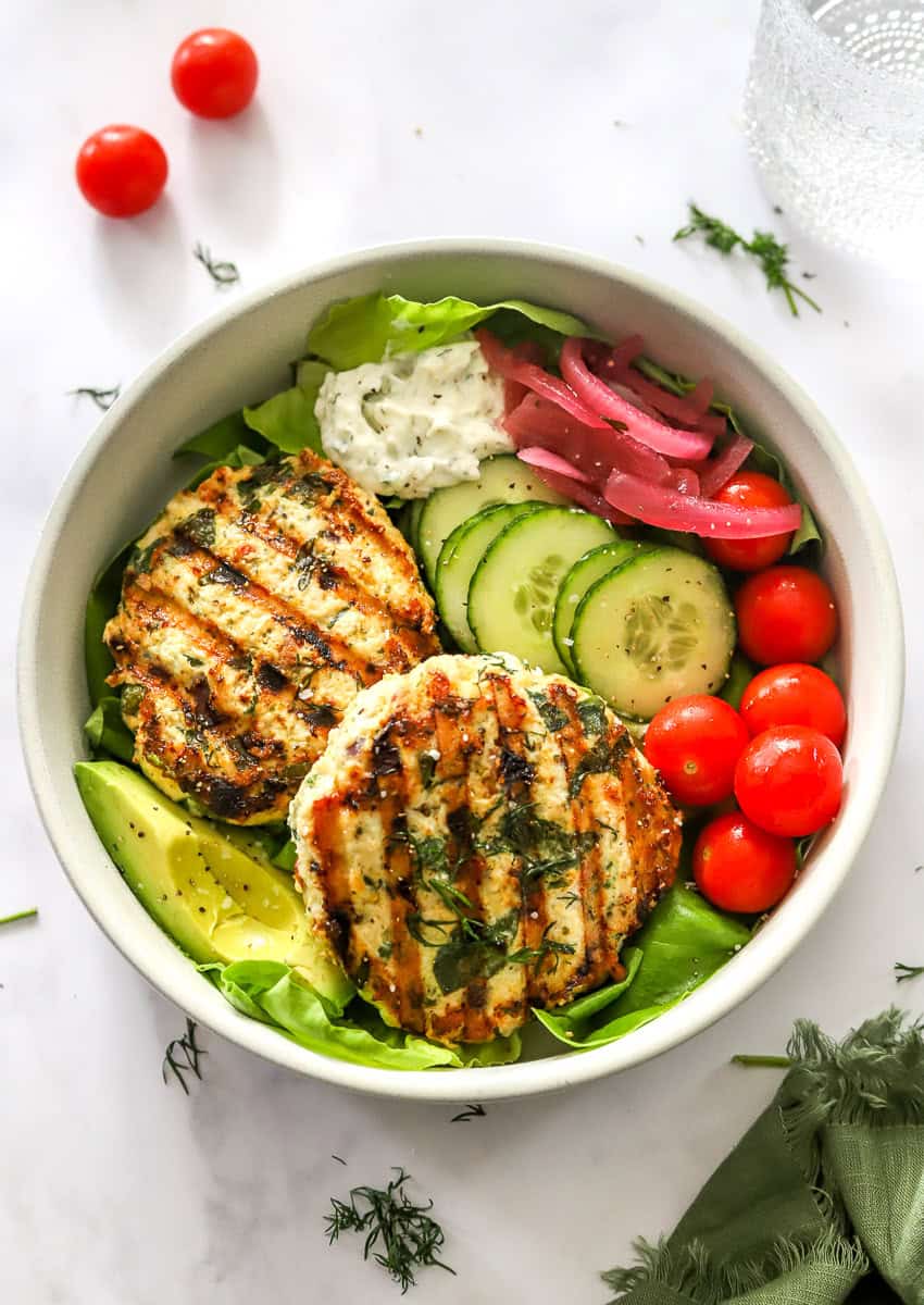 Round bowl with 2 chicken burgers, sliced cucumber, cherry tomatoes and sliced avocado in. the bowl.