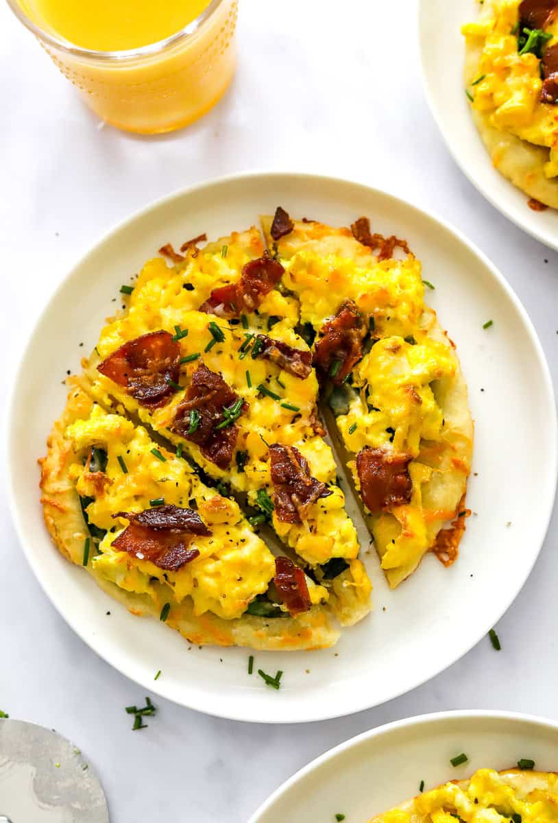 Breakfast pizza with cooked eggs and bacon cut into slices on a round white plate with another plate of it behind it and infant of it.