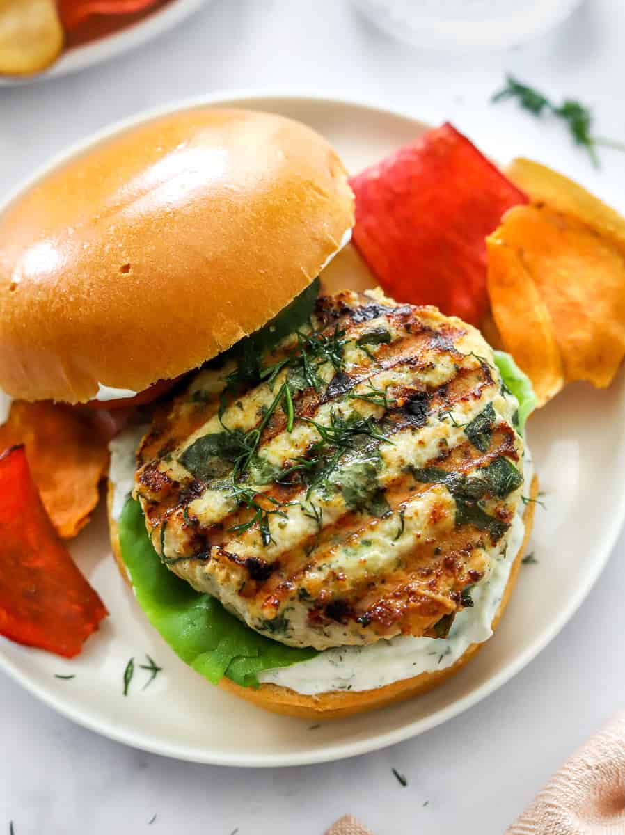 Burger bun topped with white sauce and a greek chicken burger with chips around it.