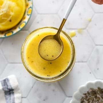 Hand lifting spoon out of jar of dijon dressing with a bowl of cut lemons, bowl of salt and pepper, and a kitchen towel around it.