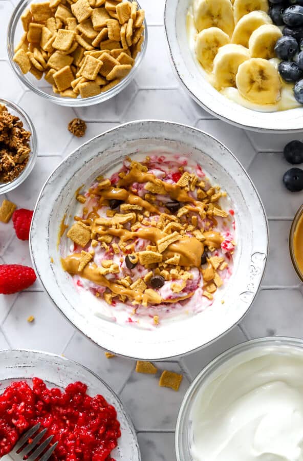 Bowl of berry flavored yogurt topped with protein cereal and nut butter with more yogurt, nut butter and mashed berries in bowls around it.