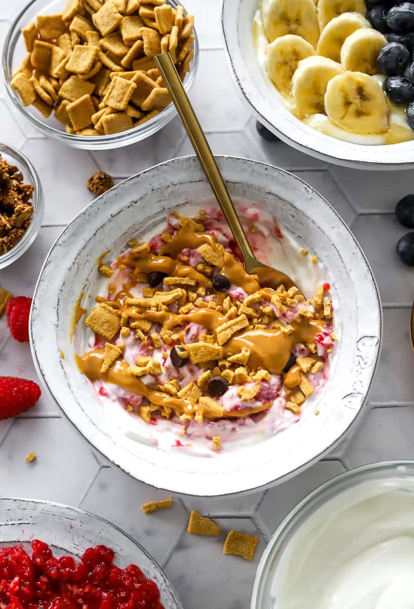 Bowl with raspberry yogurt topped with crunchy cereal and nut butter with more berries, cereal, fruit and yogurt in bowls around it.