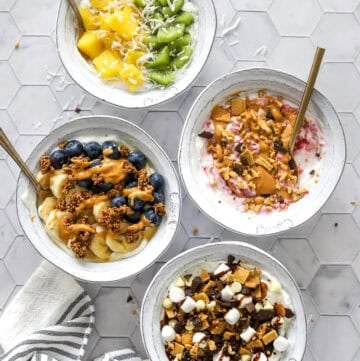 4 Greek yogurt bowls with various toppings with spoons in the bowls and a striped towel in front of it.