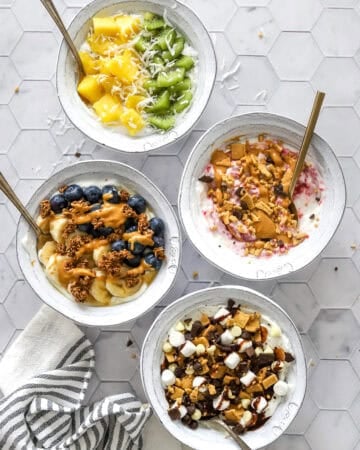 4 Greek yogurt bowls with various toppings with spoons in the bowls and a striped towel in front of it.