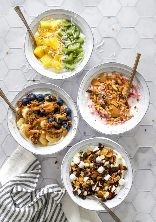 4 Greek yogurt bowls with various toppings with spoons in the bowls and a striped towel in front of it.