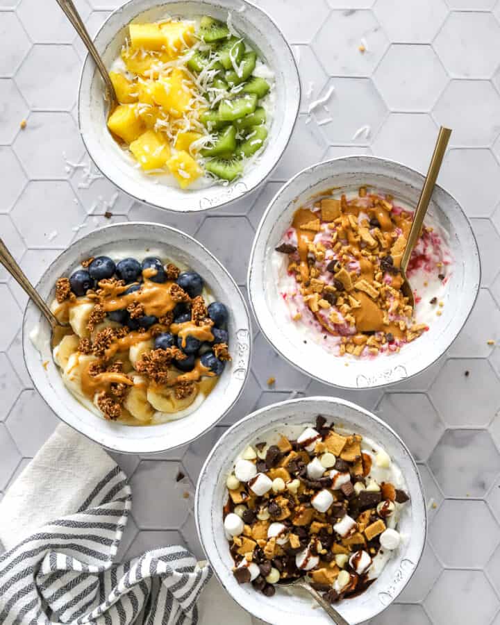 4 Greek yogurt bowls with various toppings with spoons in the bowls and a striped towel in front of it.