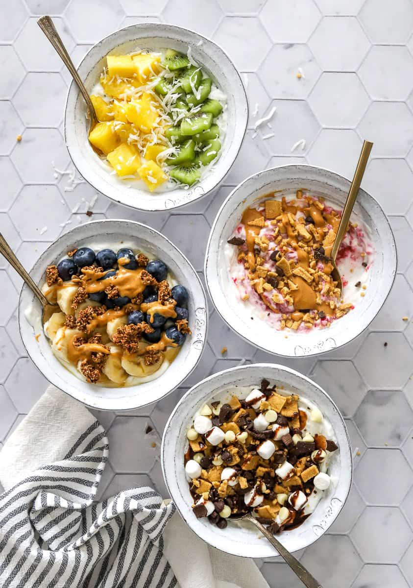 4 Greek yogurt bowls with various toppings with spoons in the bowls and a striped towel in front of it.