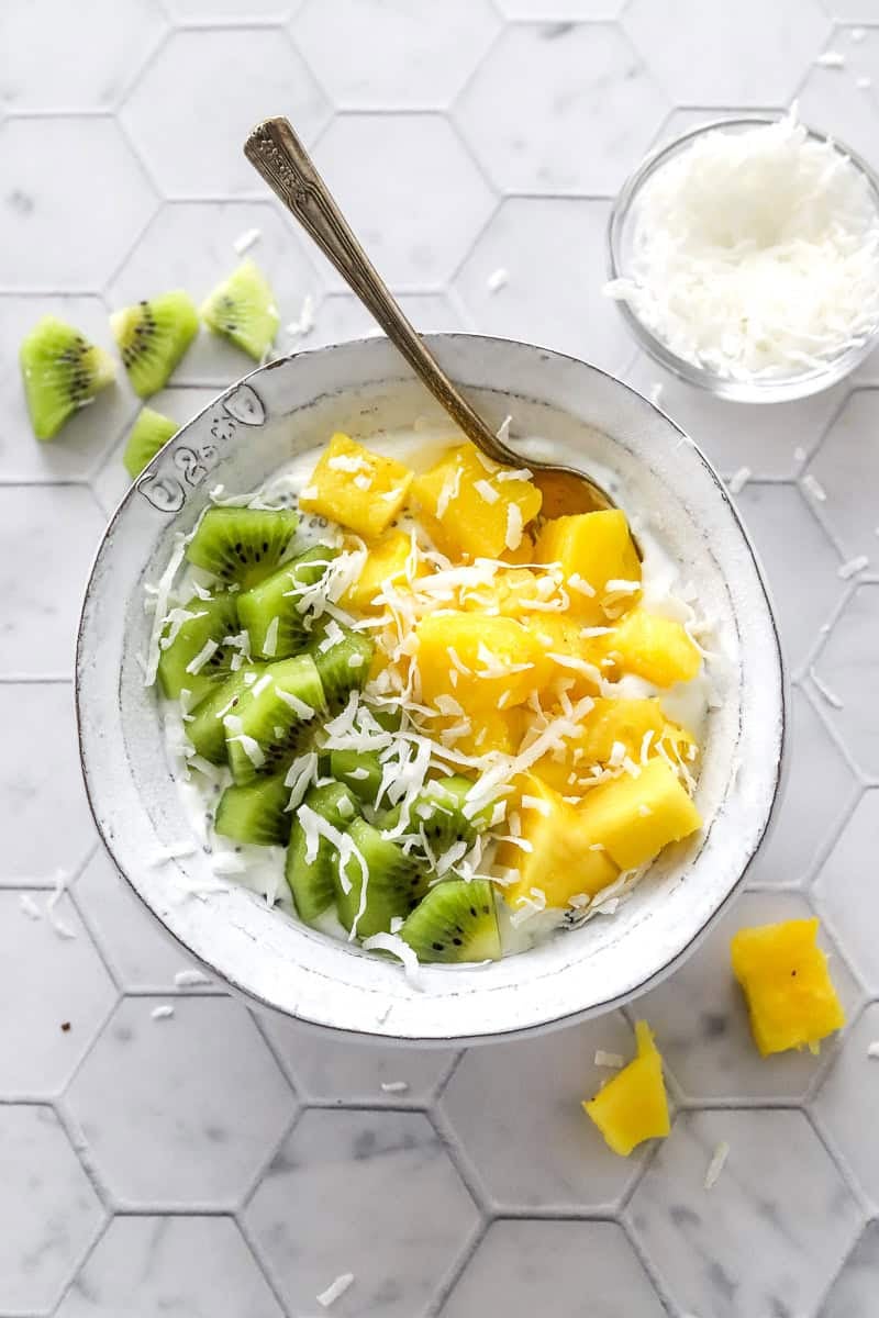Greek yogurt topped with chopped kiwi and pineapple, topped with shredded coconut in a bowl with a spoon in the bowl.