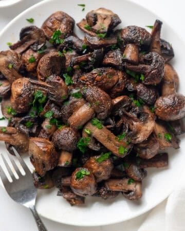 Plate of air fryer mushrooms with a fork on the plate and bowl of parsley behind it.