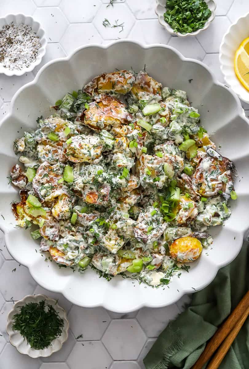Crispy smashed potato salad mixed in a serving bowl with more herbs and lemon slices in bowls around it.