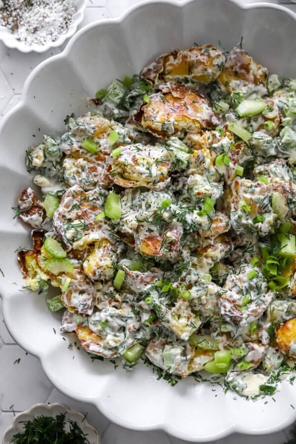 Crispy potatoes mixed with celery herbs and creamy yogurt dressing in a salad bowl with a bowl of herbs in front of it.
