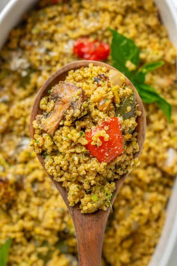 Quinao tossed in green sauce with bell peppers on a wood spoon with the casserole behind it.