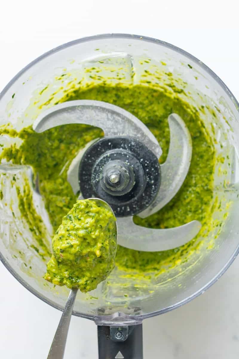 Spoon holding up blended green pesto with more in a food processor behind it.