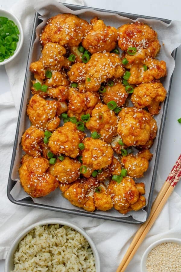 Baked breaded cauliflower florets on baking pan with cooked rice and chopsticks in front of it.