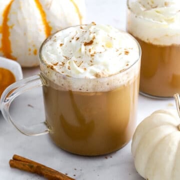 Fall pumpkin spice latte recipe with whipped cream in see through mugs with white pumpkins and cinnamon sticks in front of them.