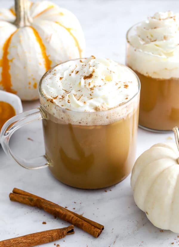 Fall pumpkin spice latte recipe with whipped cream in see through mugs with white pumpkins and cinnamon sticks in front of them.