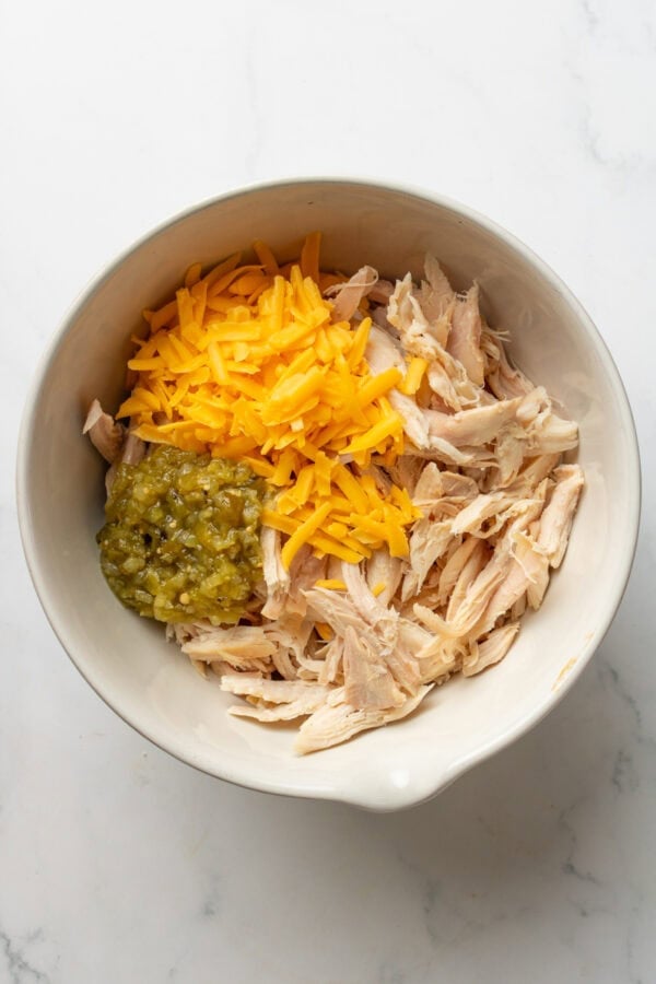Bowl filled with cooked shredded chicken, salsa verde, and shredded cheddar cheese.