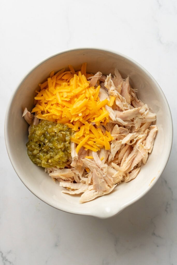 Bowl filled with cooked shredded chicken, salsa verde, and shredded cheddar cheese.