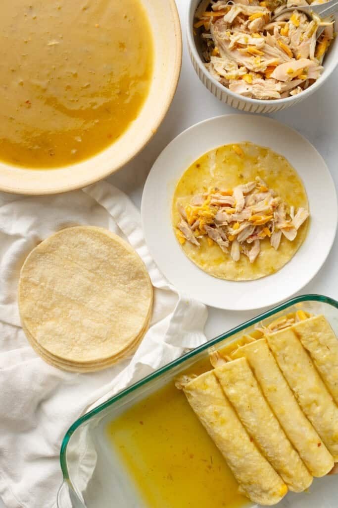 Chicken enchiladas assembly station with a glass pan of assembled enchiladas, with a stack of corn tortillas, plate with tortilla with chicken and cheese on it.