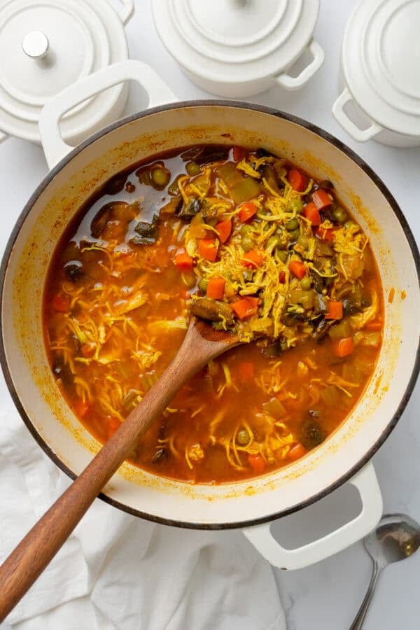 Best healthy chicken soup in a white pot with a wood spoon with some small white pots with lids behind it.