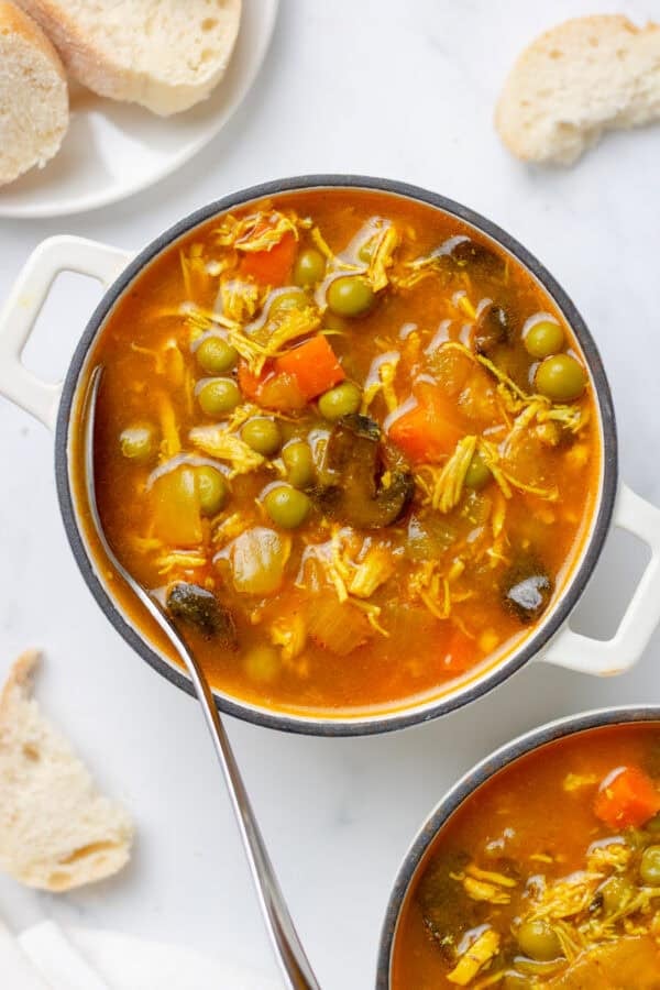 Bowl of healthy chicken soup with a spoon in it with bread behind it and another bowl of soup in front of it.