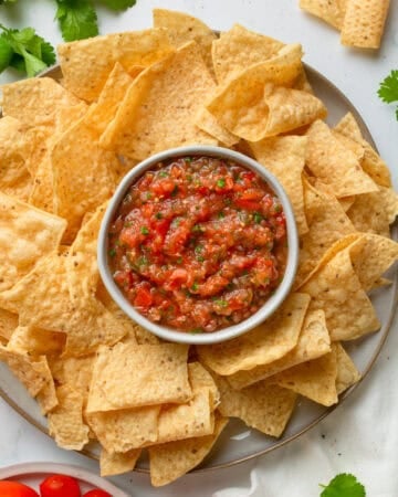 Tomato salas in a bowl on a platter with tortilla chips around it.