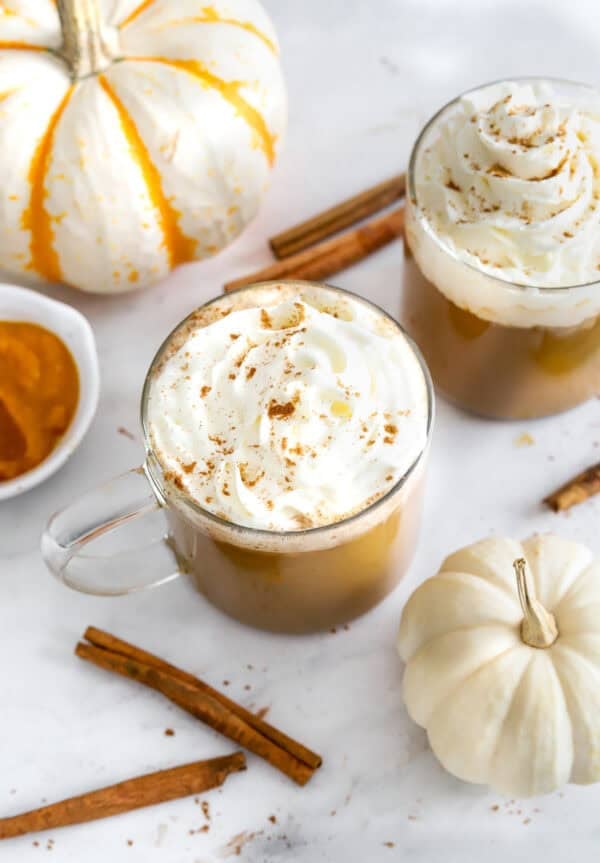 Latte's in glass mugs with whipped cream on them and white pumpkins around them.