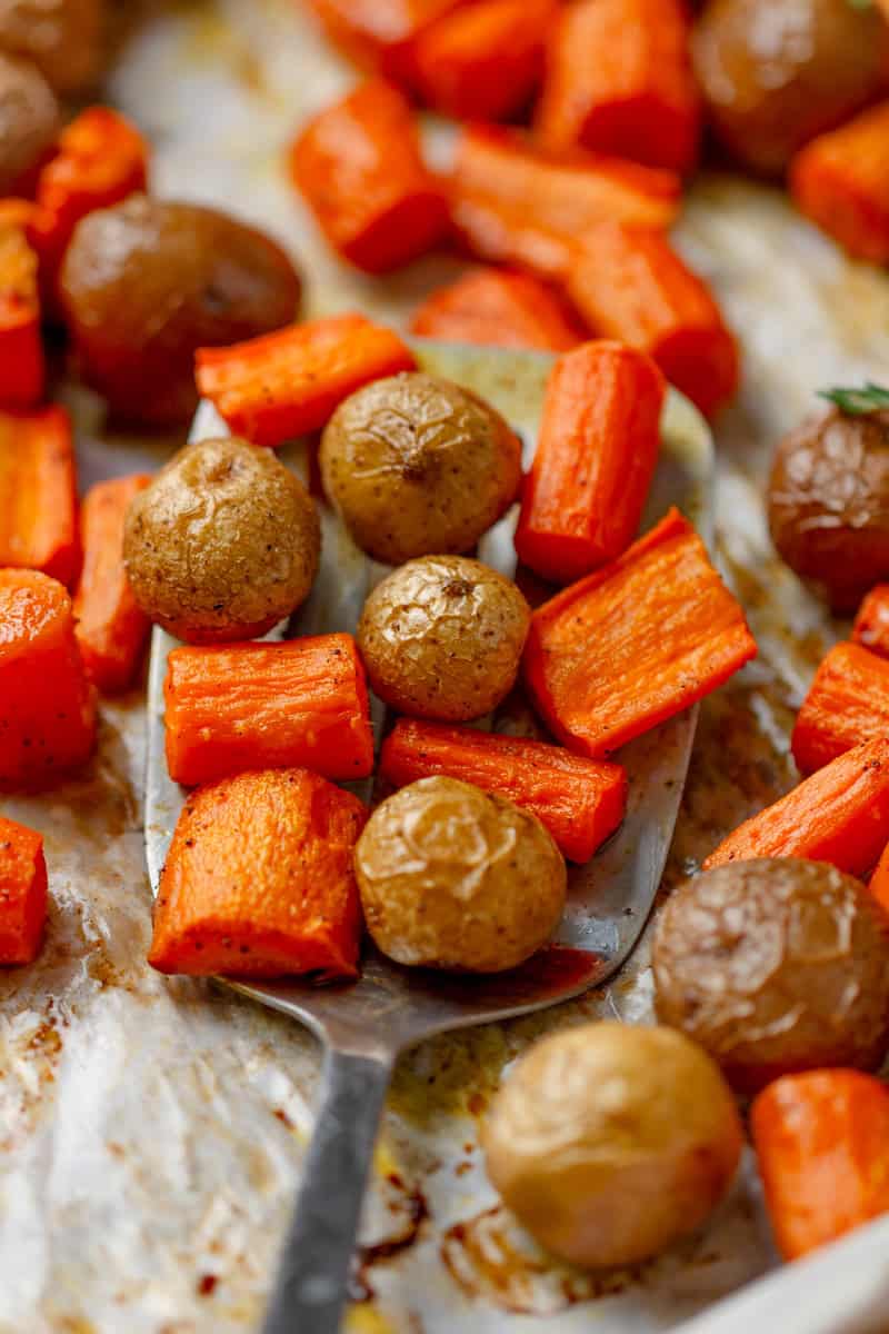 Cooked carrots and potatoes on a baking pan with a silver spatula on the pan with some on it.