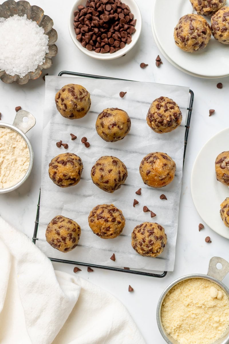 Chocolate chips edible cookie ought bites with on a lined baking rack with more ingredients in bowls around it. 